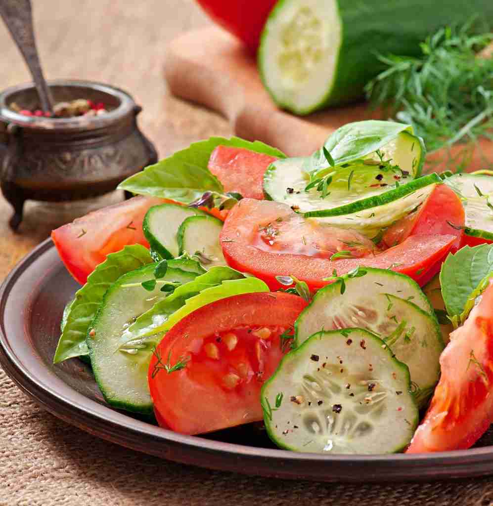 A fresh salad featuring sliced tomatoes and cucumbers sits on a wooden plate, garnished with herbs and black pepper. In the background, a cutting board holds more vegetables.