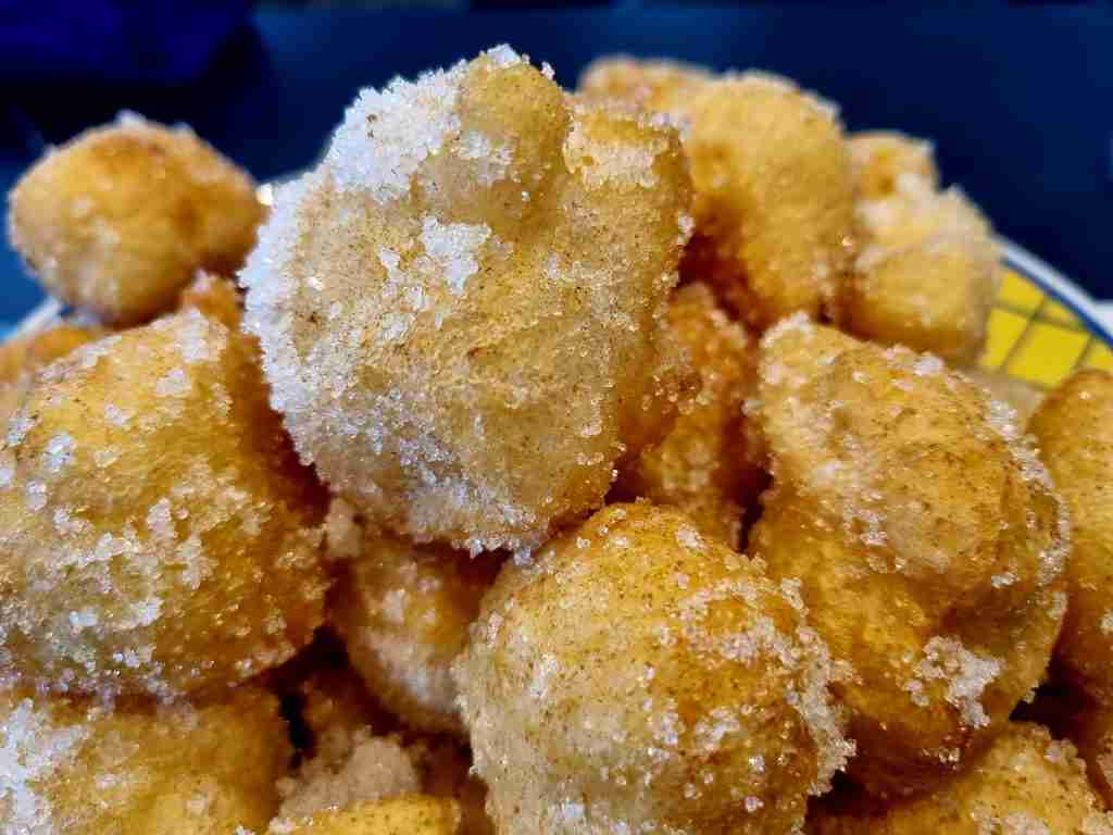 Golden-brown fritters coated in granulated sugar are piled on a plate, creating an appetizing display against a dark blue background.