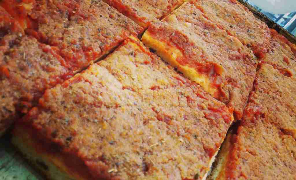 Rectangular slices of thick focaccia topped with a layer of tomato and breadcrumbs rest on a baking tray, showcasing a crispy, golden-brown surface.
